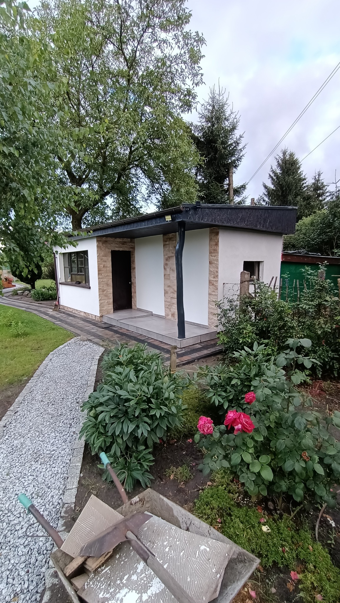 Wykończony budynek gospodarczy z elewacją imitującą kamień, widok z ogrodu z taczką wypełnioną płytkami w pierwszym planie.