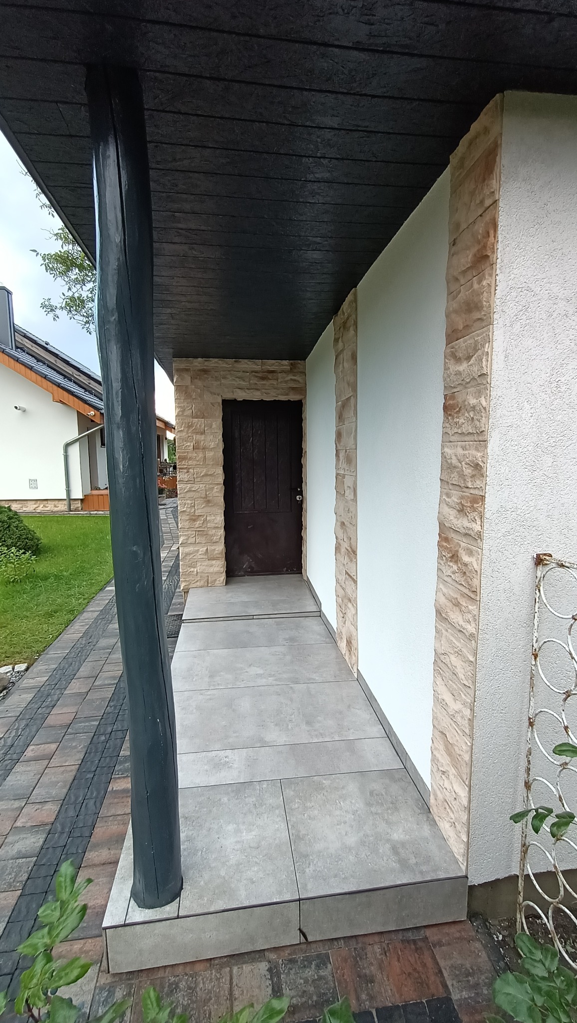 Zadaszone wejście do budynku z szarą, drewnopodobną kolumną, jasnoszarą podłogą z płytek, drzwiami w kolorze ciemnego brązu i elewacją w odcieniach bieli i piaskowca.