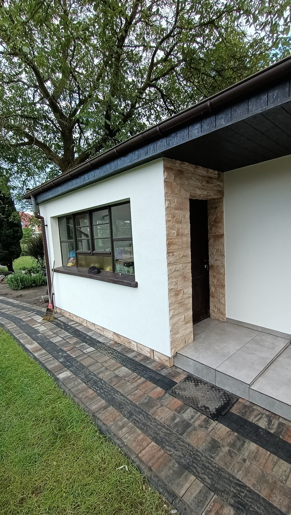 Wejście do budynku z elewacją w kolorze białym i obramowaniem drzwi z imitacji kamienia, widoczna kostka brukowa w odcieniach brązu i czerni oraz trawnik.