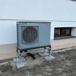 Kamil Krzystanek - Szara pompa ciepła powietrze-woda zamontowana na metalowej konstrukcji przy ścianie budynku z dwoma oknami piwnicznymi, widoczne betonowe podstawy i czarna rura odprowadzająca skropliny.
