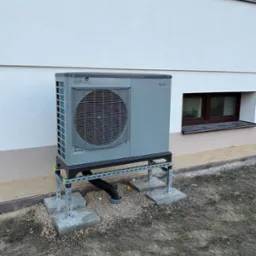 Szara pompa ciepła powietrze-woda zamontowana na metalowej konstrukcji przy ścianie budynku z dwoma oknami piwnicznymi, widoczne betonowe podstawy i czarna rura odprowadzająca skropliny.