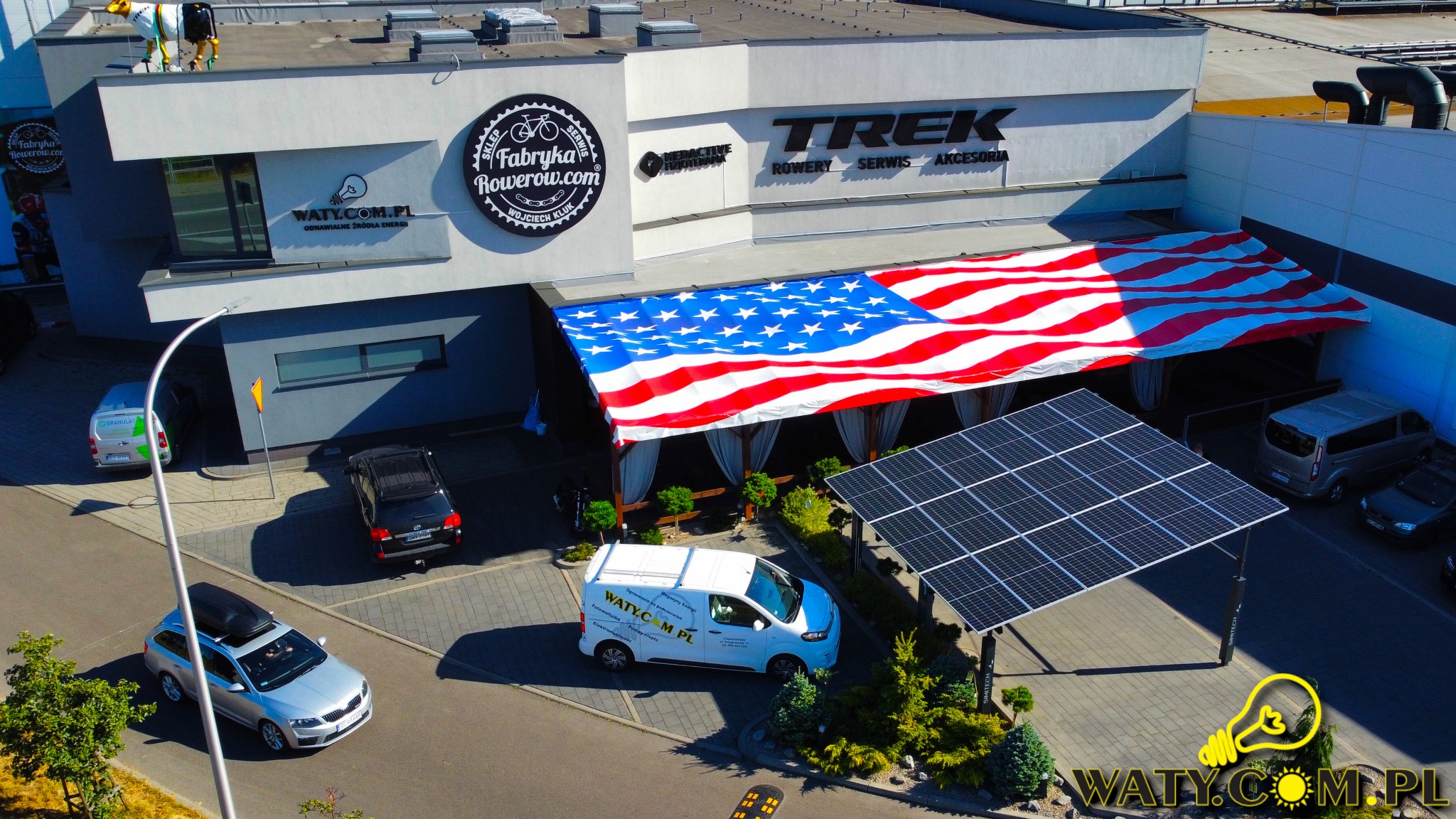 Widok z góry na budynek firmy z zainstalowanymi panelami słonecznymi jako zadaszenie parkingu, z flagą USA nad wejściem i zaparkowanymi samochodami.