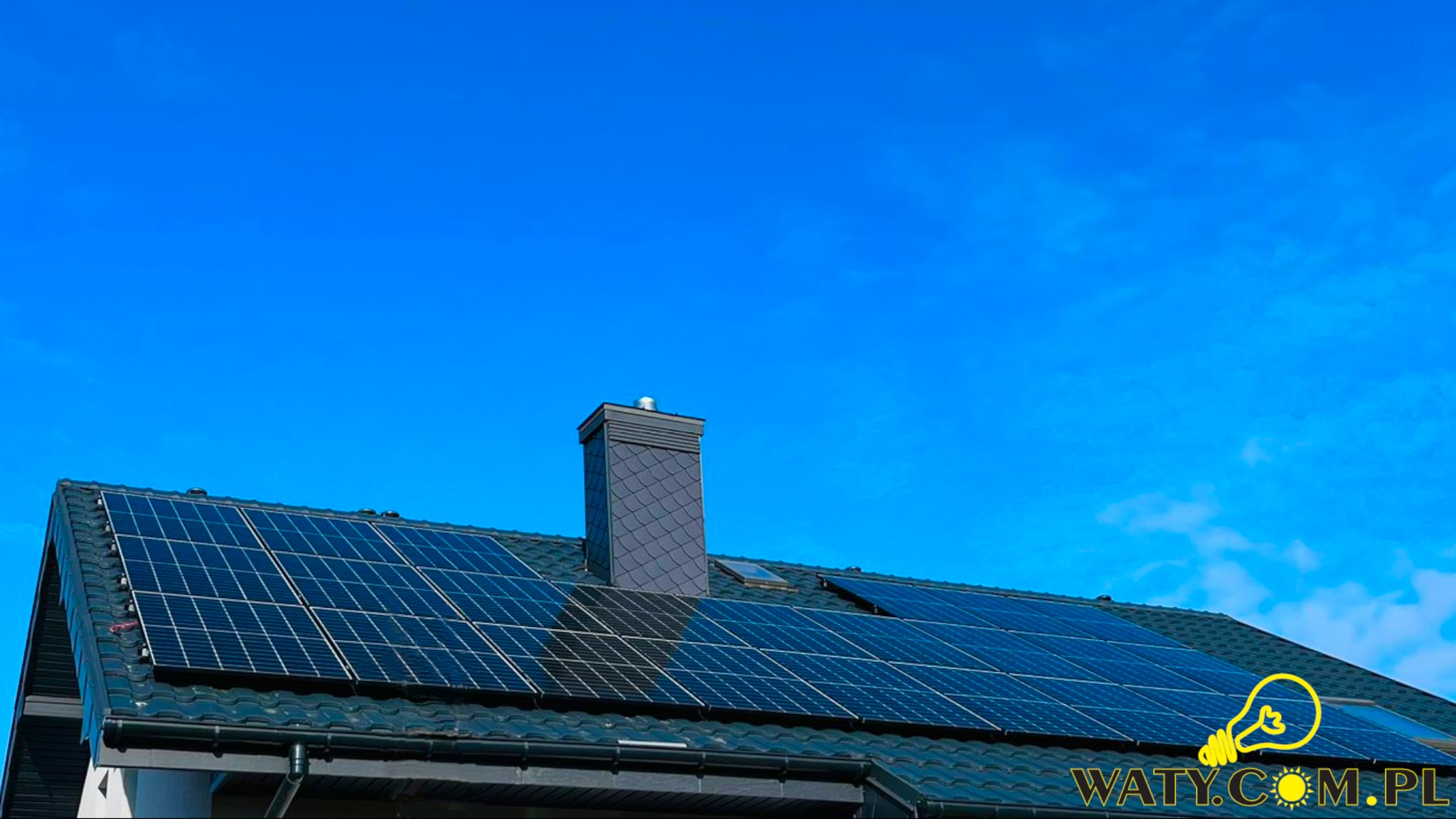 Panele fotowoltaiczne na ciemnym dachu domu jednorodzinnego, widoczne także komin z szarej dachówki i błękitne niebo z delikatnymi chmurami.