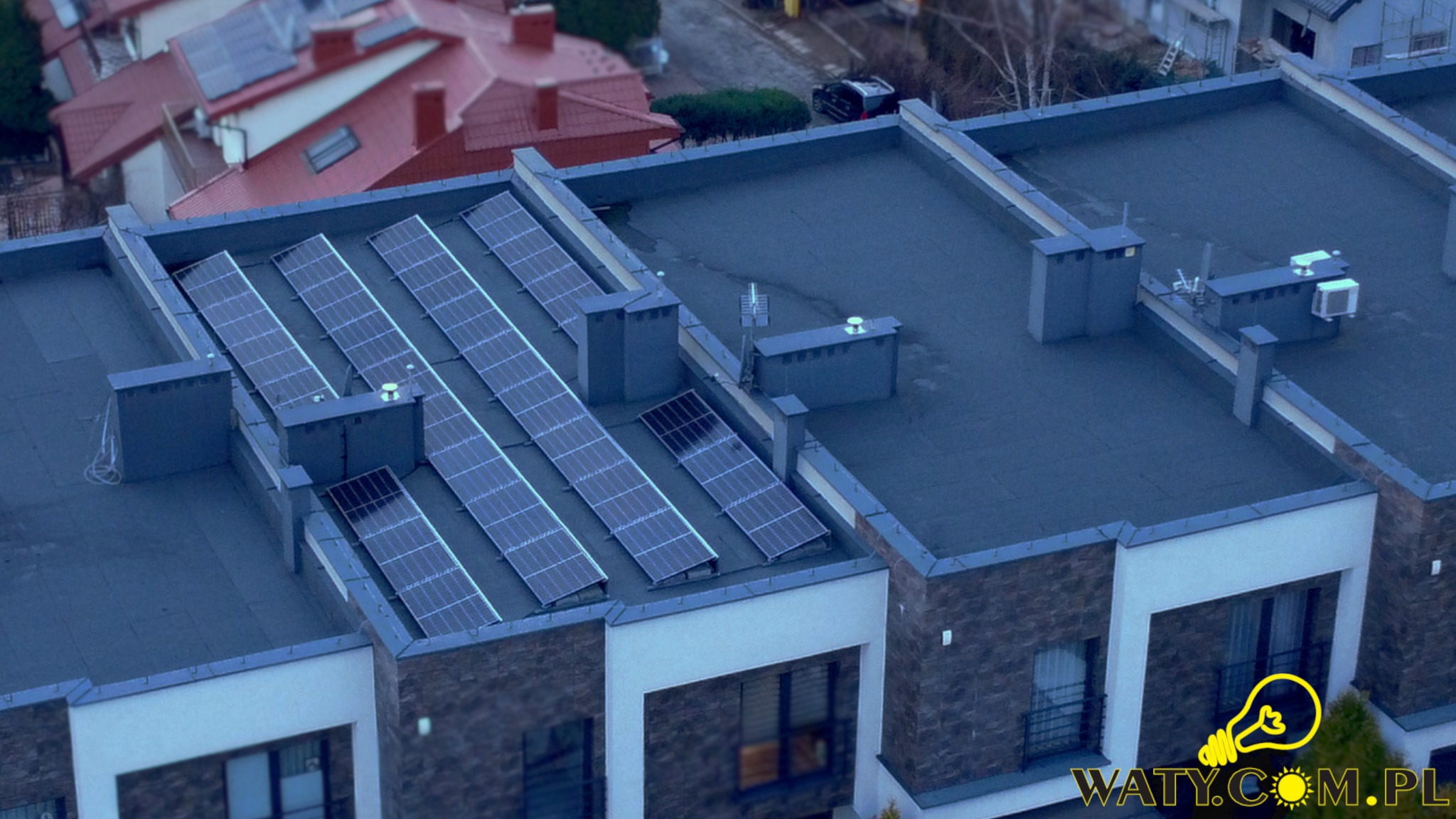 Widok z góry na dach budynku z zainstalowanymi panelami fotowoltaicznymi i klimatyzatorami, w tle inne budynki mieszkalne.