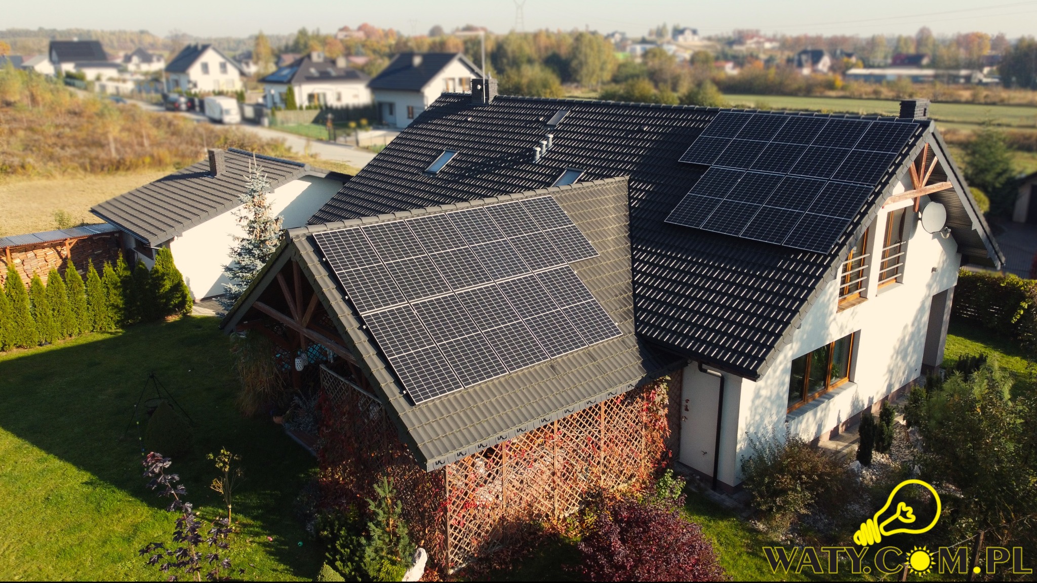 Dom jednorodzinny z panelami słonecznymi na dachu pokrytym ciemną dachówką, widok z góry, jesienne otoczenie z drzewami w tle.