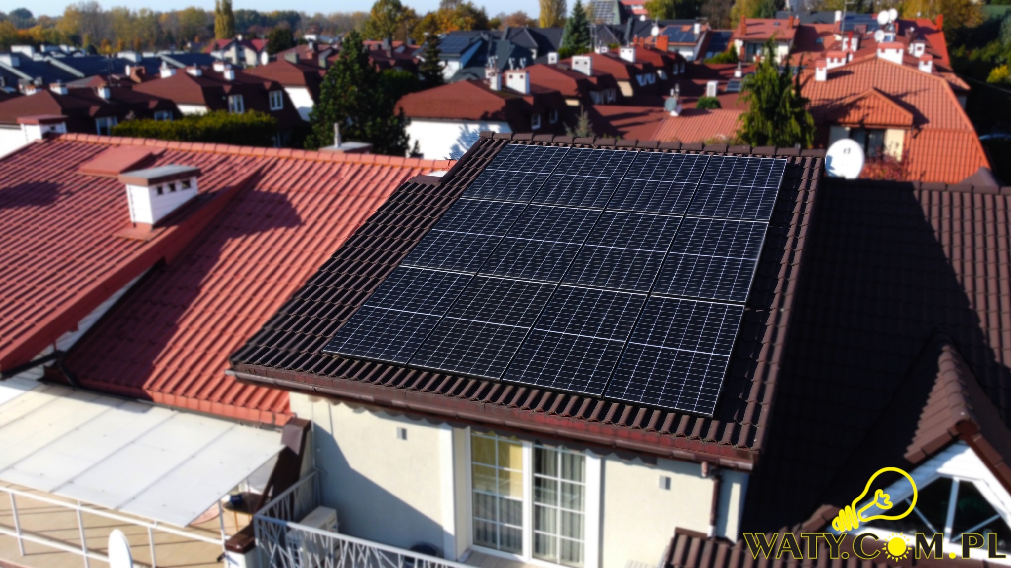 Panele słoneczne na brązowym dachu z dachówki ceramicznej, widok z góry na osiedle domów jednorodzinnych z czerwonymi dachami w słoneczny dzień.