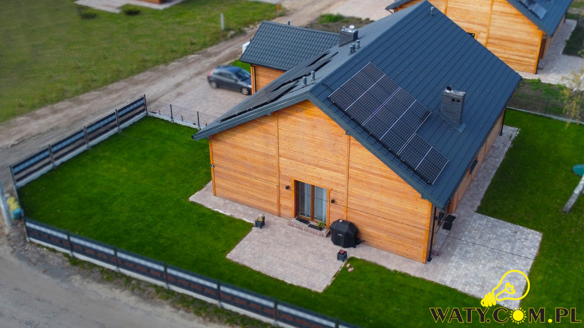 Widok z góry na drewniany dom z ciemną dachówką i panelami fotowoltaicznymi, otoczony zielonym trawnikiem i ogrodzeniem, z samochodem zaparkowanym w tle.
