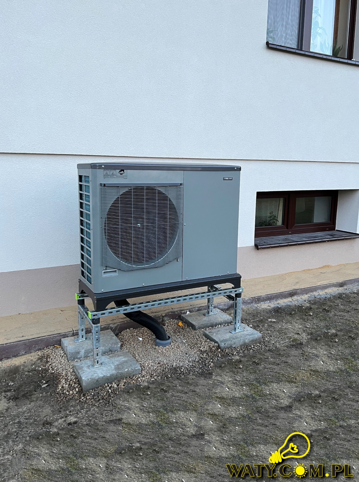 Szara pompa ciepła powietrze-woda zamontowana na metalowej konstrukcji przy ścianie budynku z dwoma oknami piwnicznymi, widoczne betonowe podstawy i czarna rura odprowadzająca skropliny.