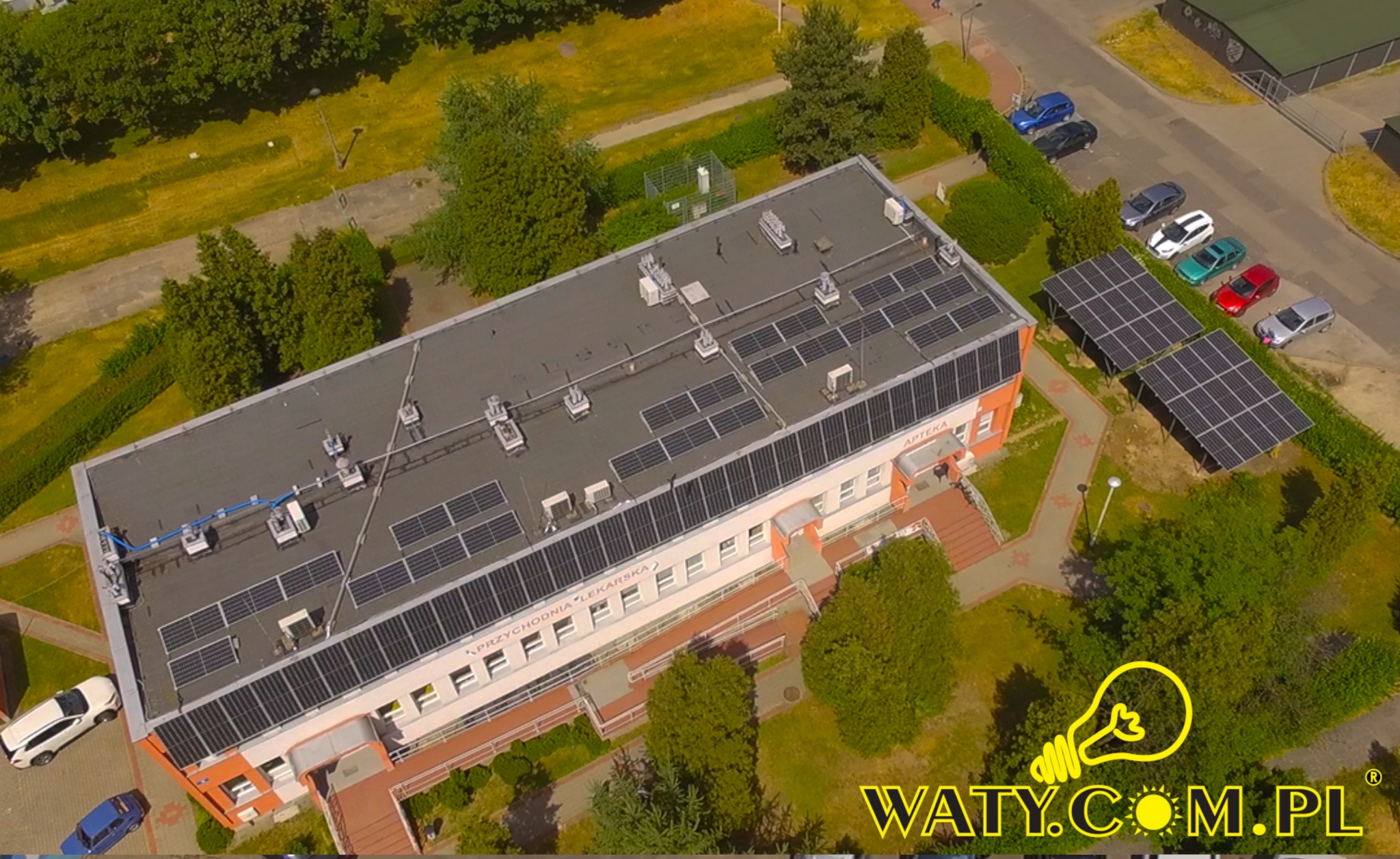 Widok z góry na budynek przychodni lekarskiej z zainstalowanymi panelami słonecznymi na dachu i obok parkingu, otoczonego zielenią.