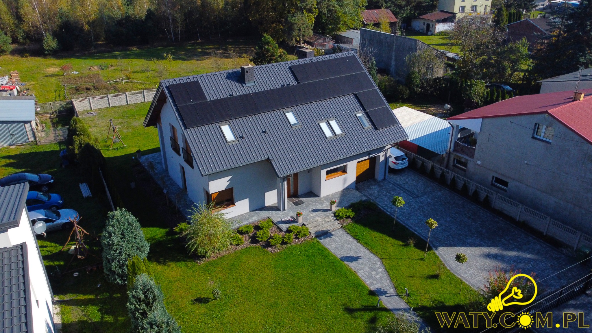 Nowoczesny dom jednorodzinny z szarym dachem i panelami fotowoltaicznymi, widok z lotu ptaka, zadbany ogród z kostką brukową i drzewami.