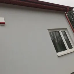 Fragment elewacji domu po malowaniu na szaro, z widocznym oknem, orynnowaniem w kolorze ceglastym i zamontowanym alarmem.