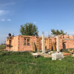 Usługi budowlane Ihor Yavorskyi - Budowa domu jednorodzinnego z czerwonej cegły, widoczne szalunki okienne, betonowe słupy i stosy bloczków na trawie, pracownik na rusztowaniu w tle.