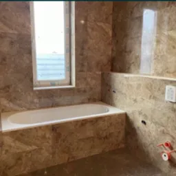 Wykończona łazienka z beżowymi marmurkowymi płytkami, białą wanną obudowaną płytkami, oknem z białymi ramami i widocznymi instalacjami hydraulicznymi przed montażem armatury.