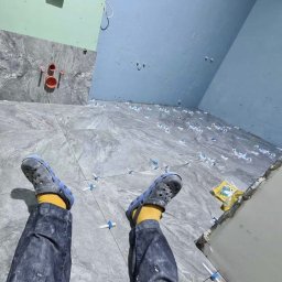 Usługi budowlane Ihor Yavorskyi - Podłoga w trakcie układania płytek imitujących marmur, widoczne krzyżyki dystansowe, rury instalacji wodnej na ścianie, niebieskie ściany i nogi wykonawcy w dżinsach i żółtych skarpetkach...