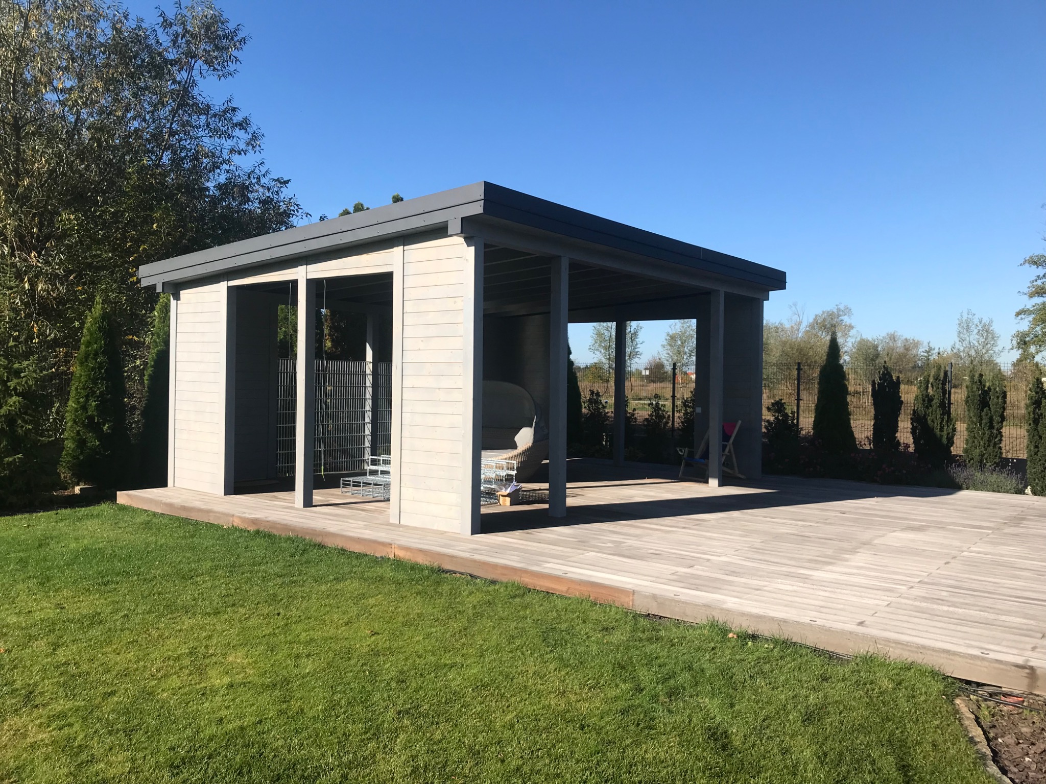 Nowoczesna altana ogrodowa z drewnianą podłogą tarasową i szarą konstrukcją, otoczona zielonym trawnikiem i tujami w słoneczny dzień.