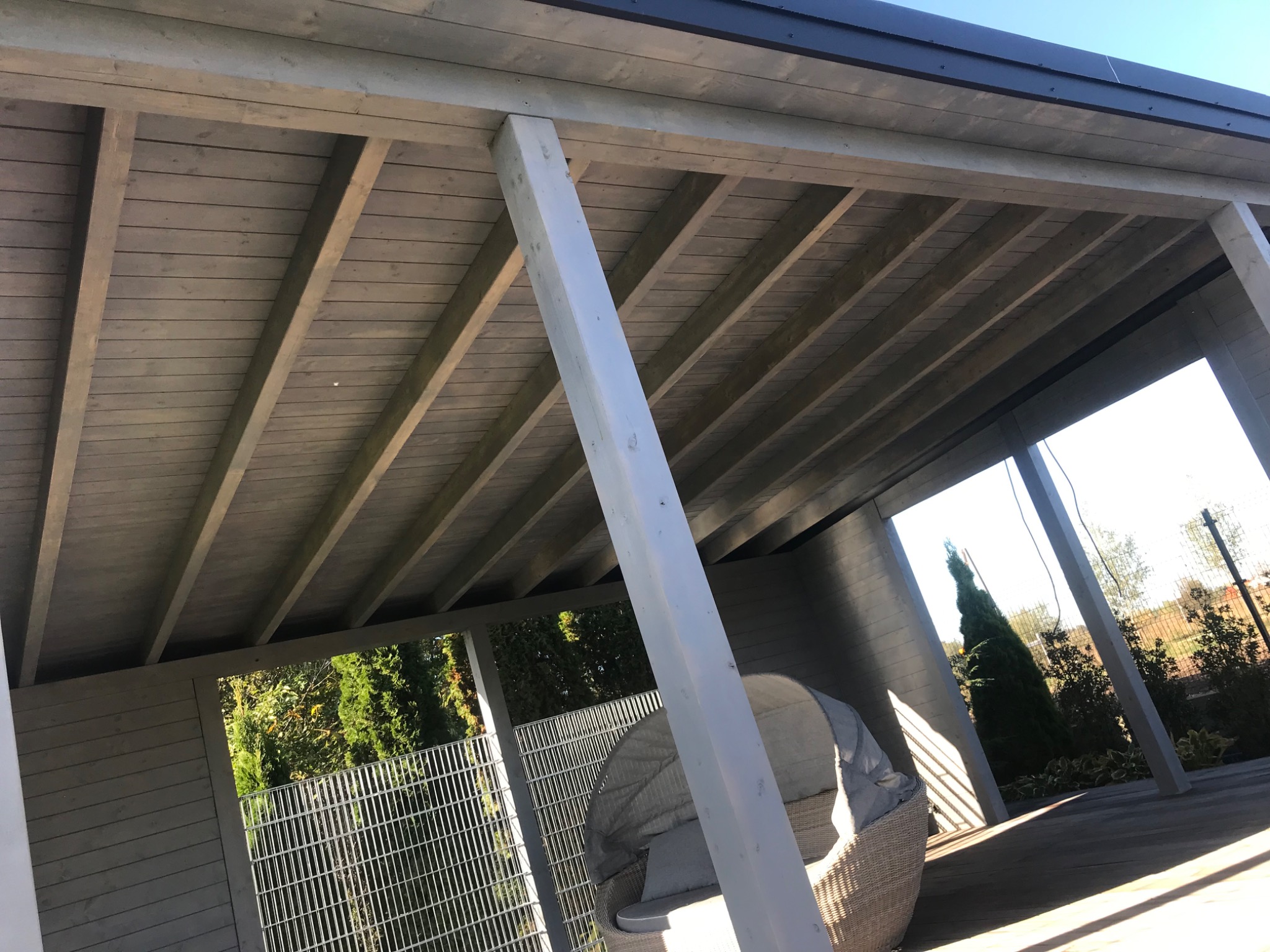 Drewniana konstrukcja z pomalowanym na szaro dachem i białymi podporami, widok z wnętrza, w tle zieleń ogrodu i krzesło z wikliny.