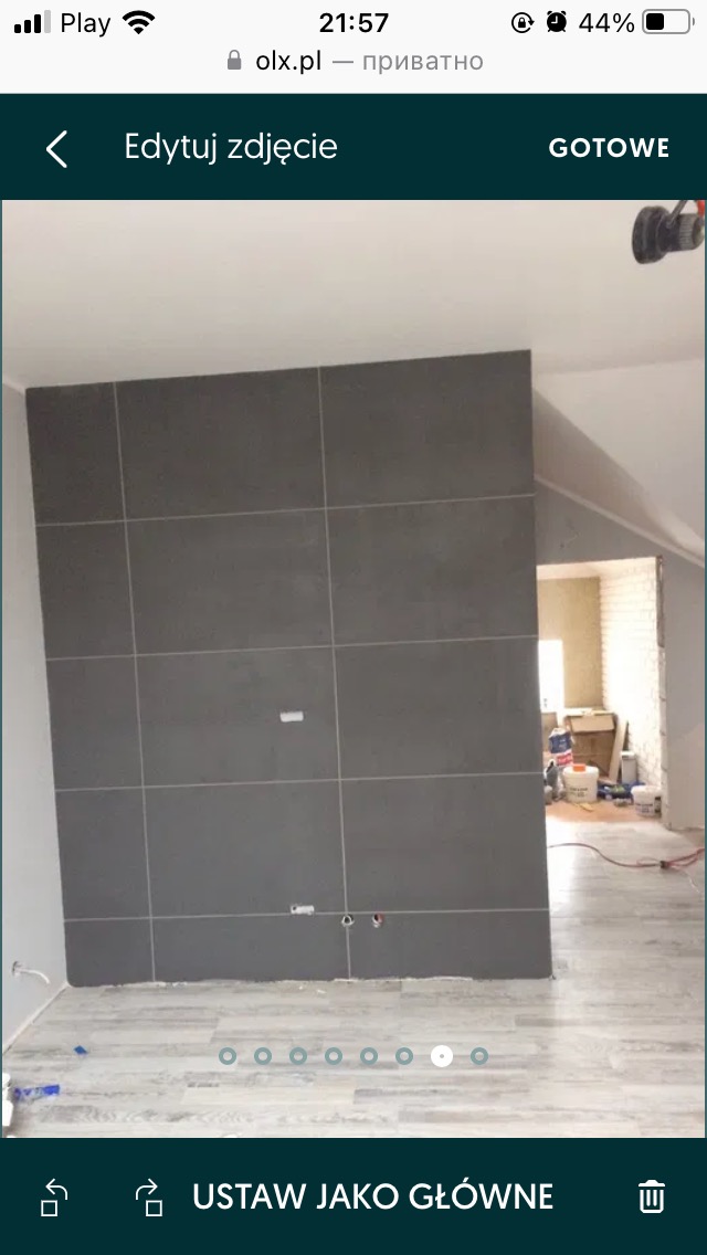 Ściana w trakcie wykańczania z szarymi płytkami, widoczne przygotowane otwory na instalacje, podłoga wyłożona panelami imitującymi drewno, w tle widoczne narzędzia i materiały budowlane.