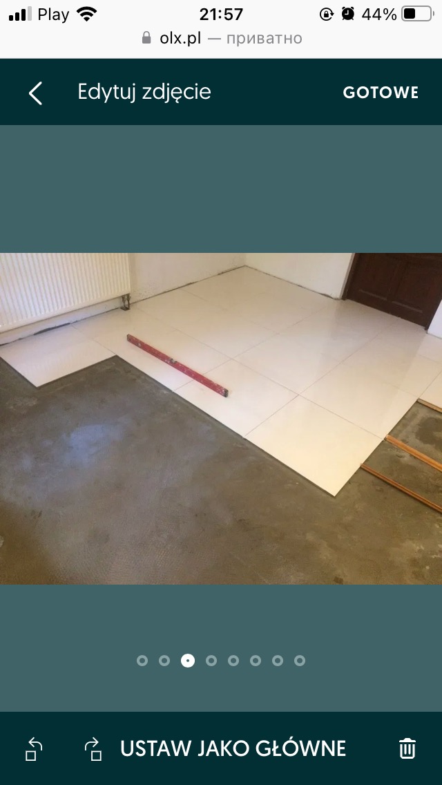 Białe płytki podłogowe układane na szarym kleju, widoczna poziomica i fragment grzejnika.