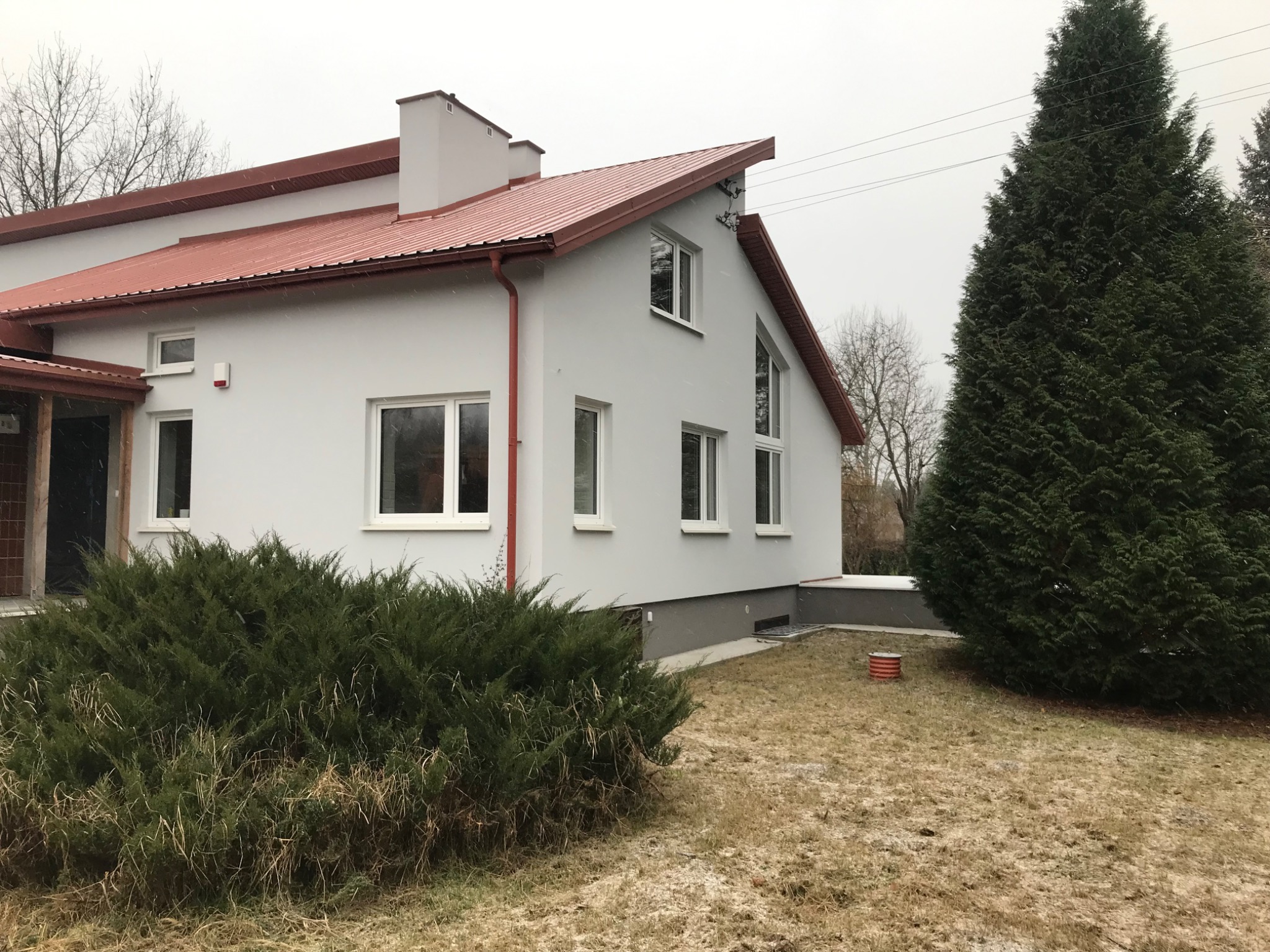 Świeżo pomalowany dom jednorodzinny z czerwoną blachodachówką, szarą elewacją i białymi ramami okiennymi, widoczny z zewnątrz w pochmurny dzień, z krzewami i drzewem iglastym na pierwszym planie.