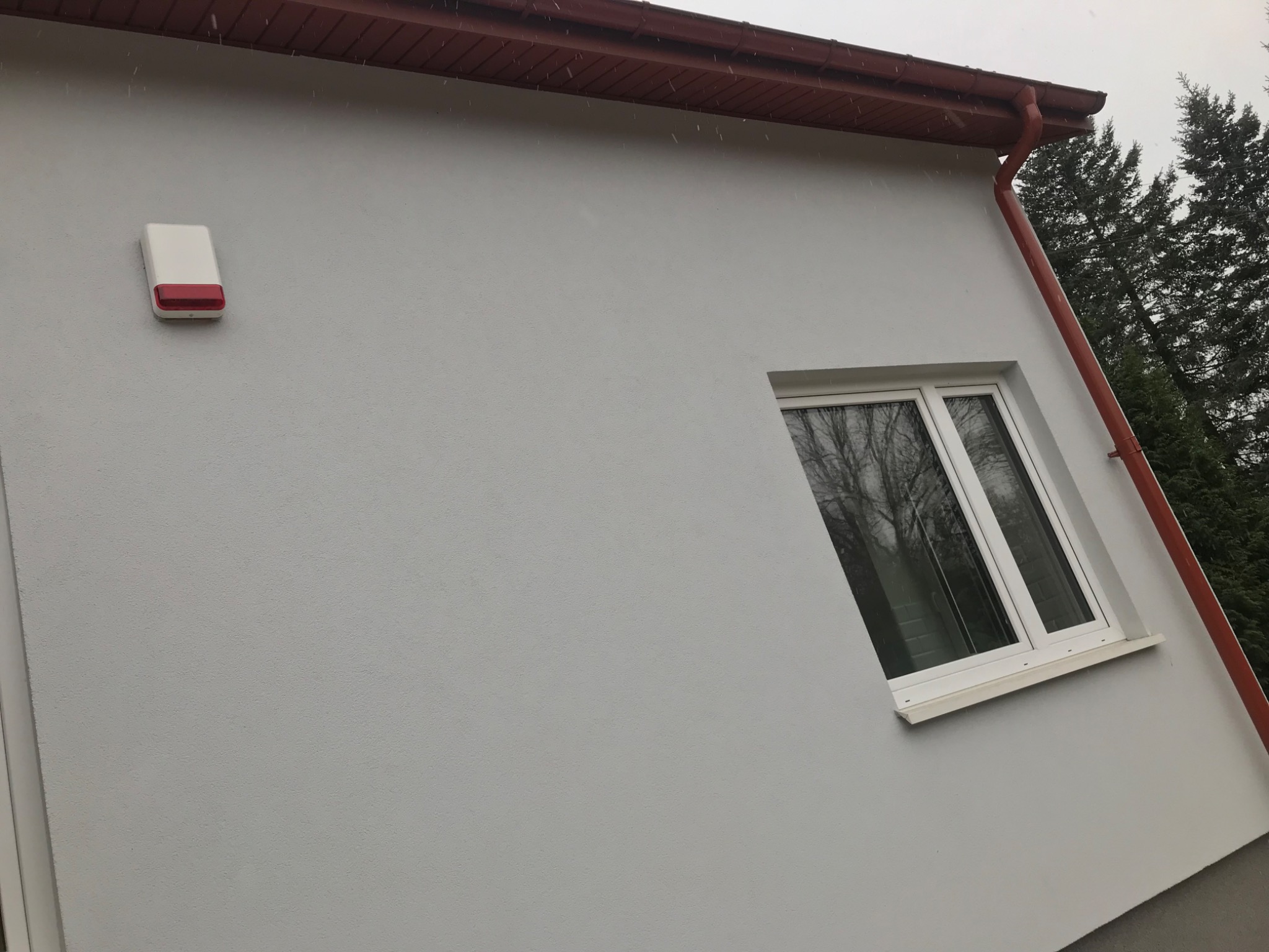 Fragment elewacji domu po malowaniu na szaro, z widocznym oknem, orynnowaniem w kolorze ceglastym i zamontowanym alarmem.
