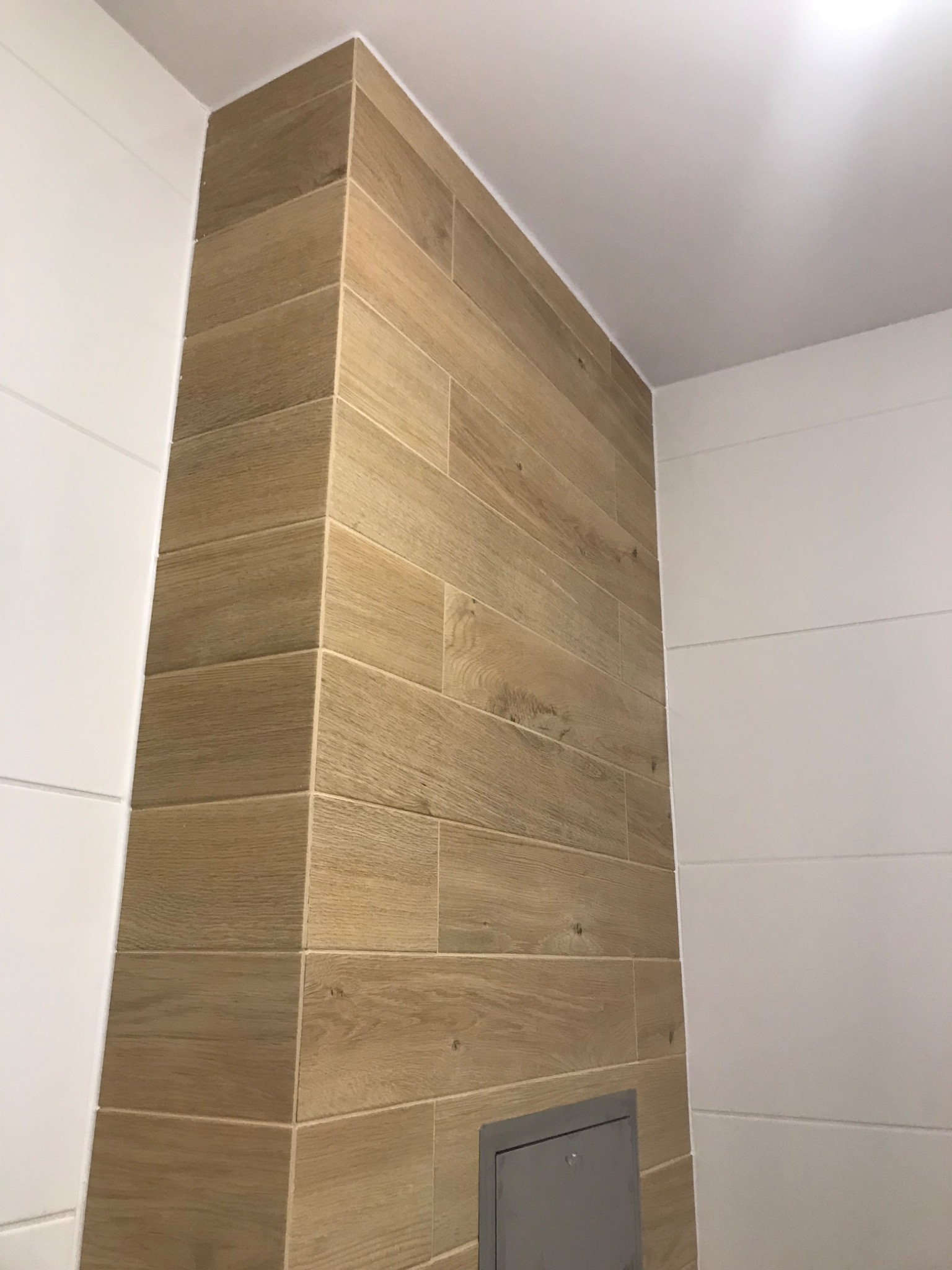 Pionowy fragment ściany w łazience wykończony płytkami imitującymi drewno, sąsiadujący z białą ścianą z poziomymi żłobieniami, widoczny szary właz rewizyjny w dolnej części.