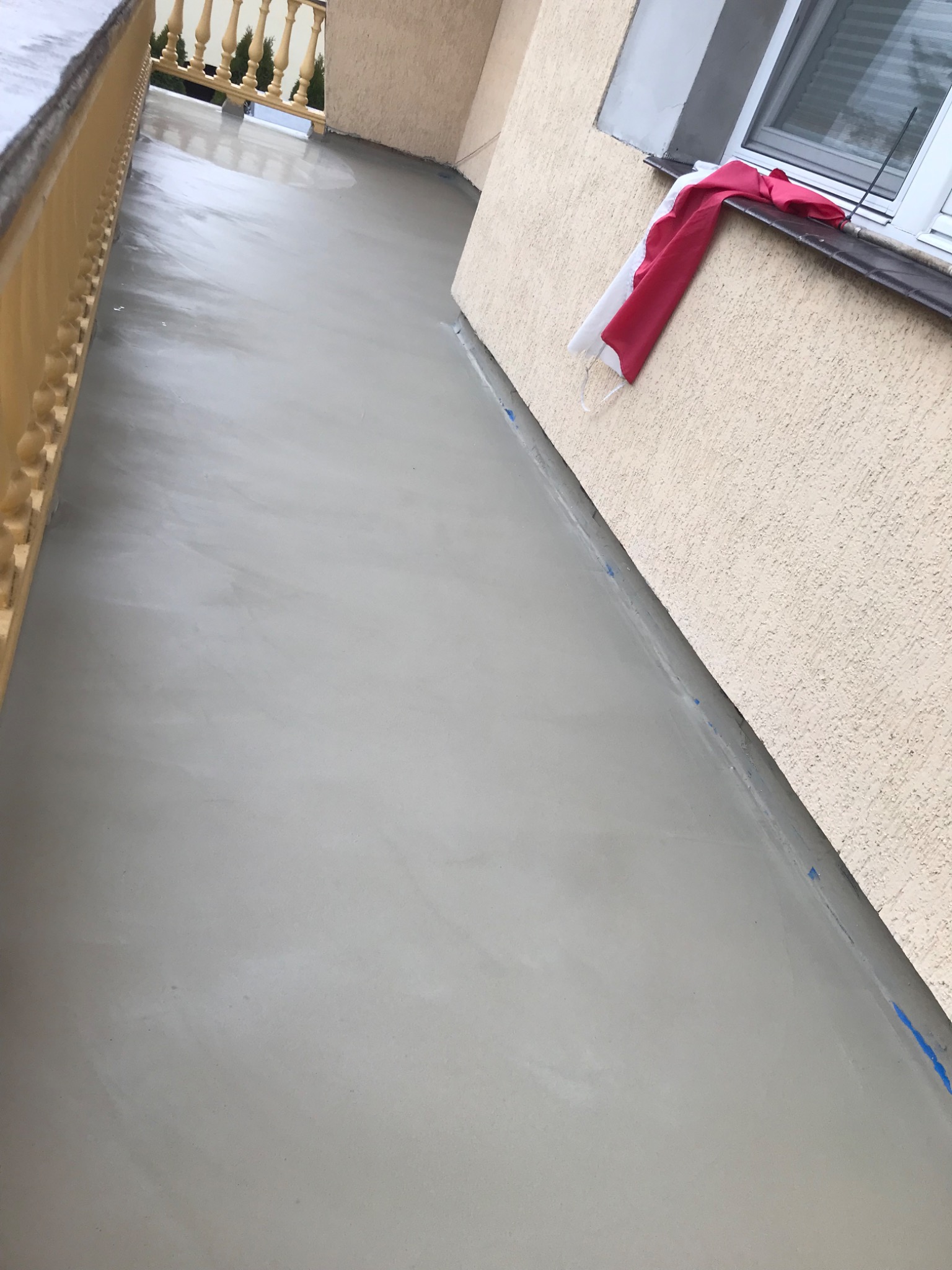 Wylany, świeży beton na balkonie w Łomiankach, zabezpieczony niebieską taśmą malarską przy ścianie. Na parapecie okna leży czerwono-biała szmatka.