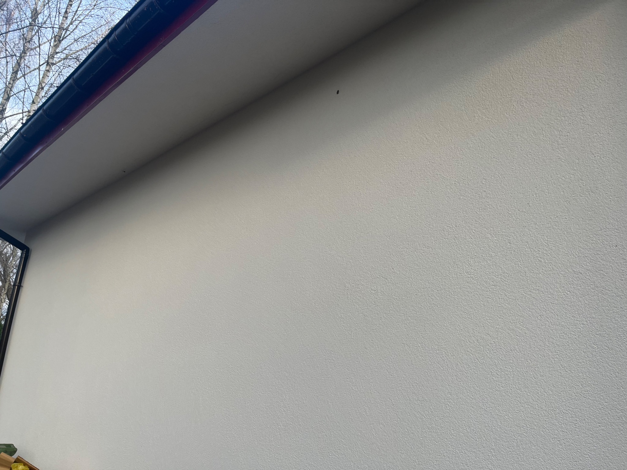 Fragment nowej, jasnej elewacji budynku z widoczną fakturą tynku i fragmentem dachu z rynnami w górnej części kadru.