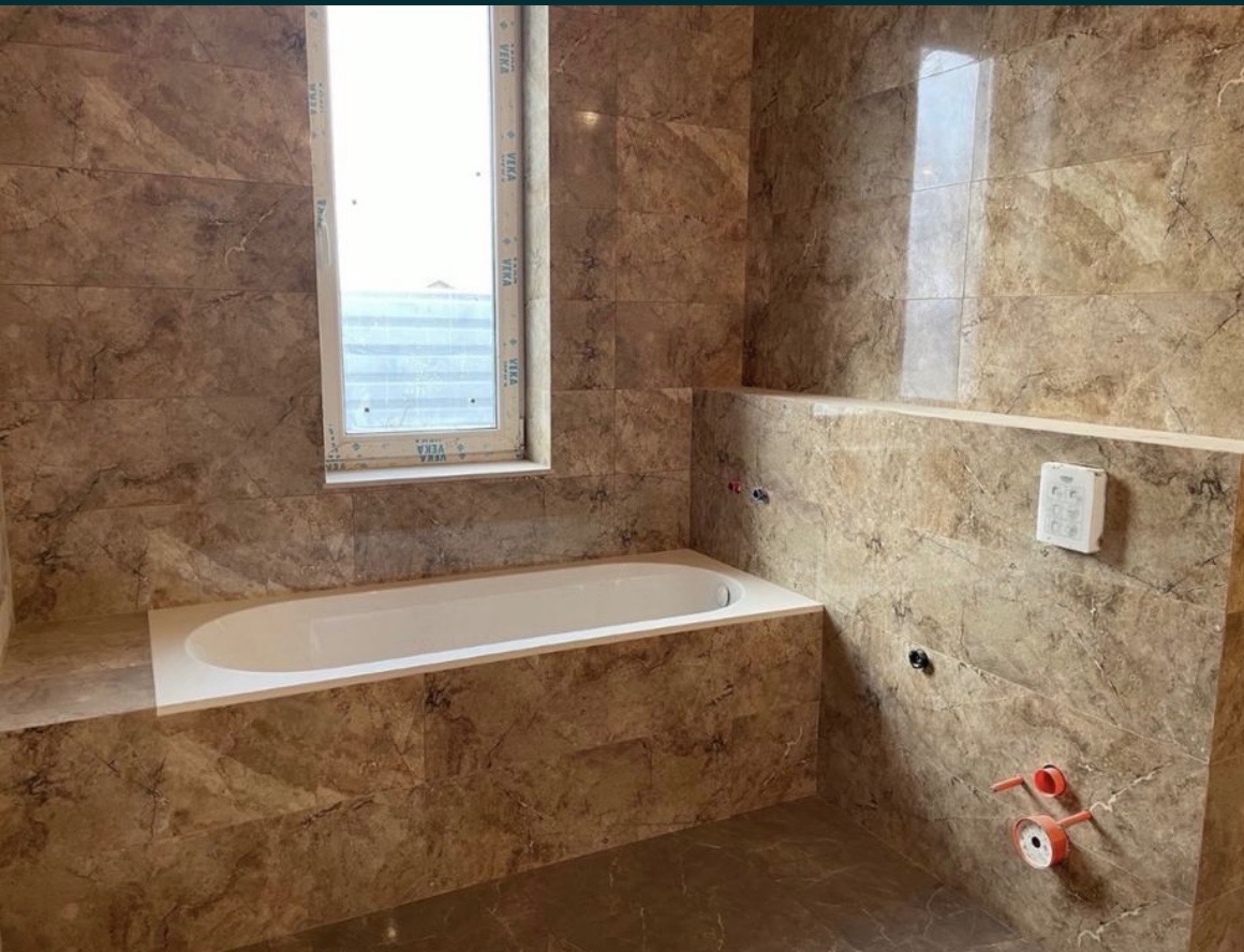 Wykończona łazienka z beżowymi marmurkowymi płytkami, białą wanną obudowaną płytkami, oknem z białymi ramami i widocznymi instalacjami hydraulicznymi przed montażem armatury.