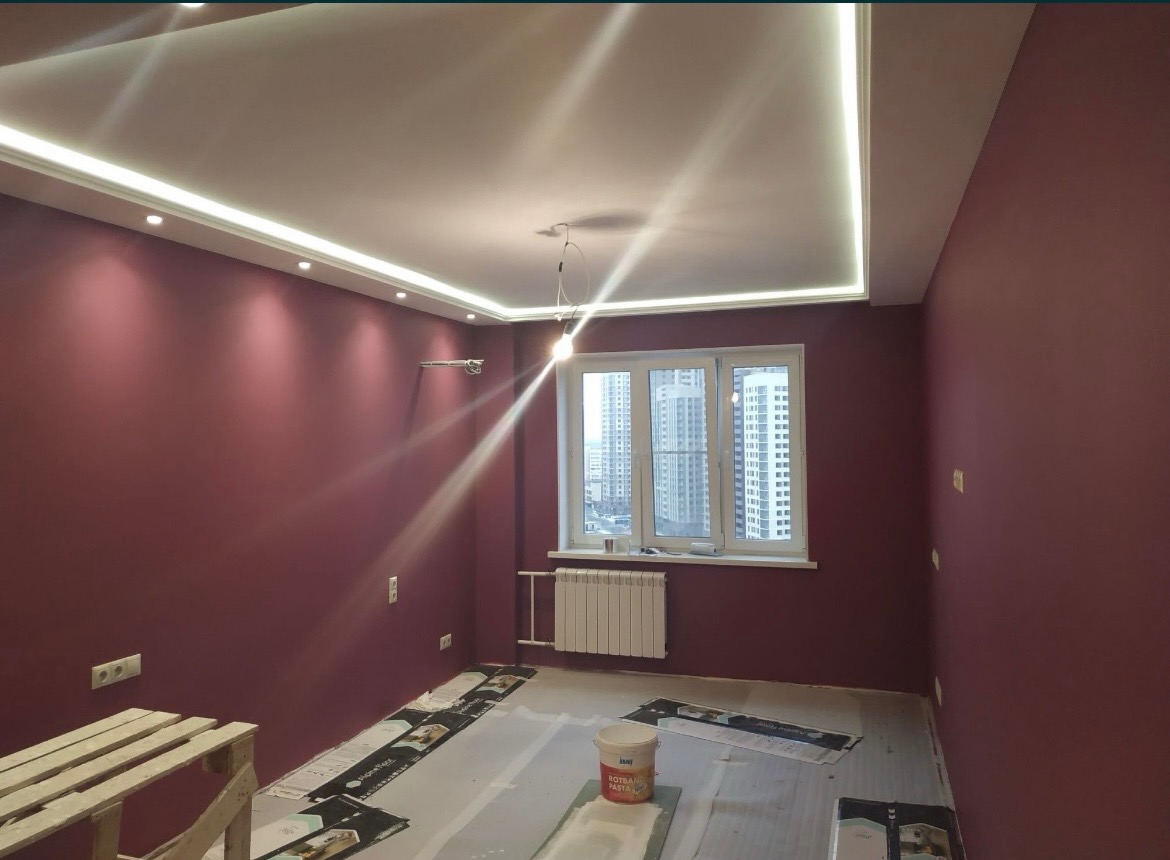 Pokój po świeżym malowaniu ścian na kolor burgundowy z białym sufitem z oświetleniem LED, widoczne zabezpieczenia podłogi i wiadro po paście gipsowej.