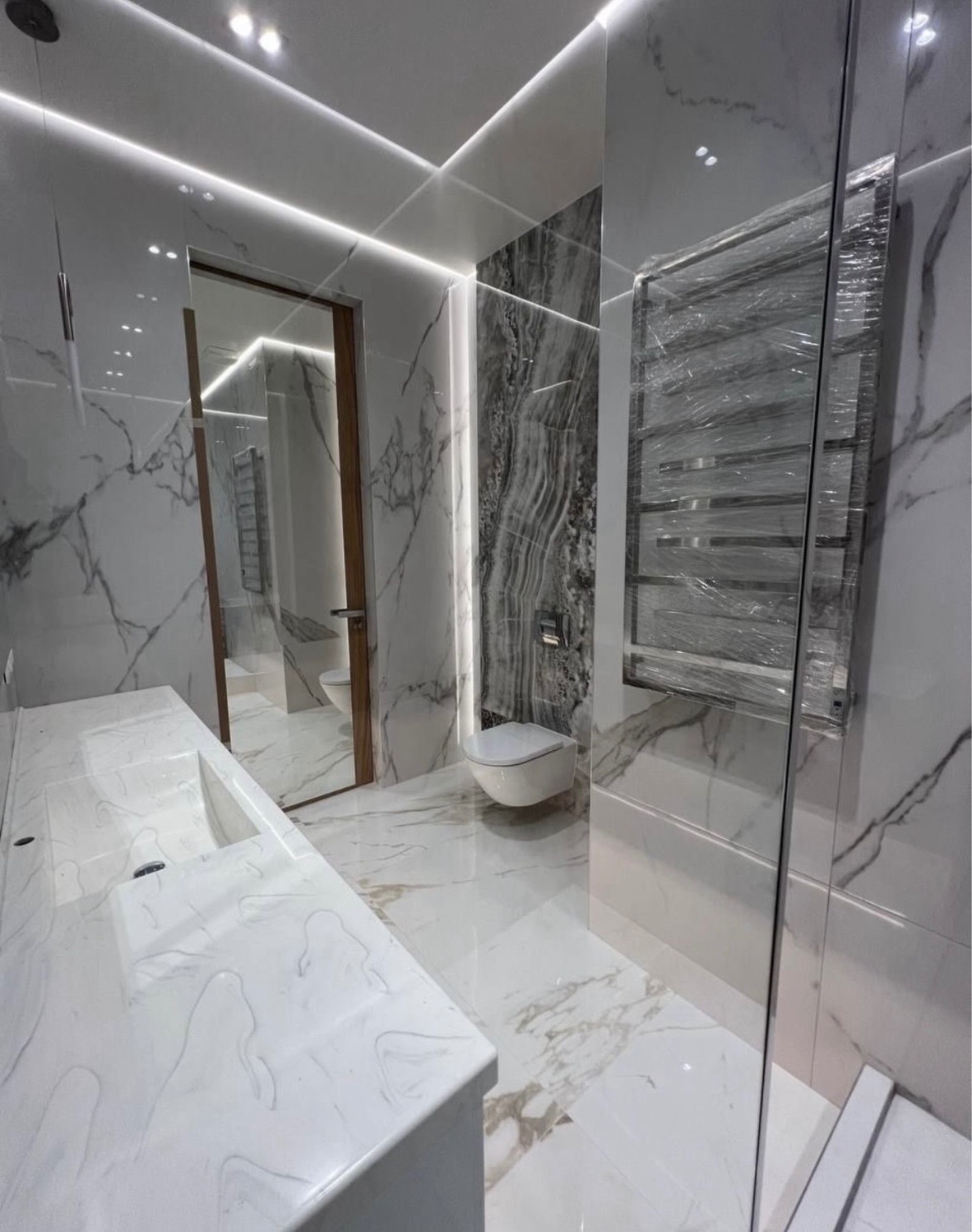 Nowoczesna łazienka wykończona białym marmurem z elementami szarości, podświetlona listwami LED, z wiszącą toaletą i grzejnikiem owiniętym folią ochronną.