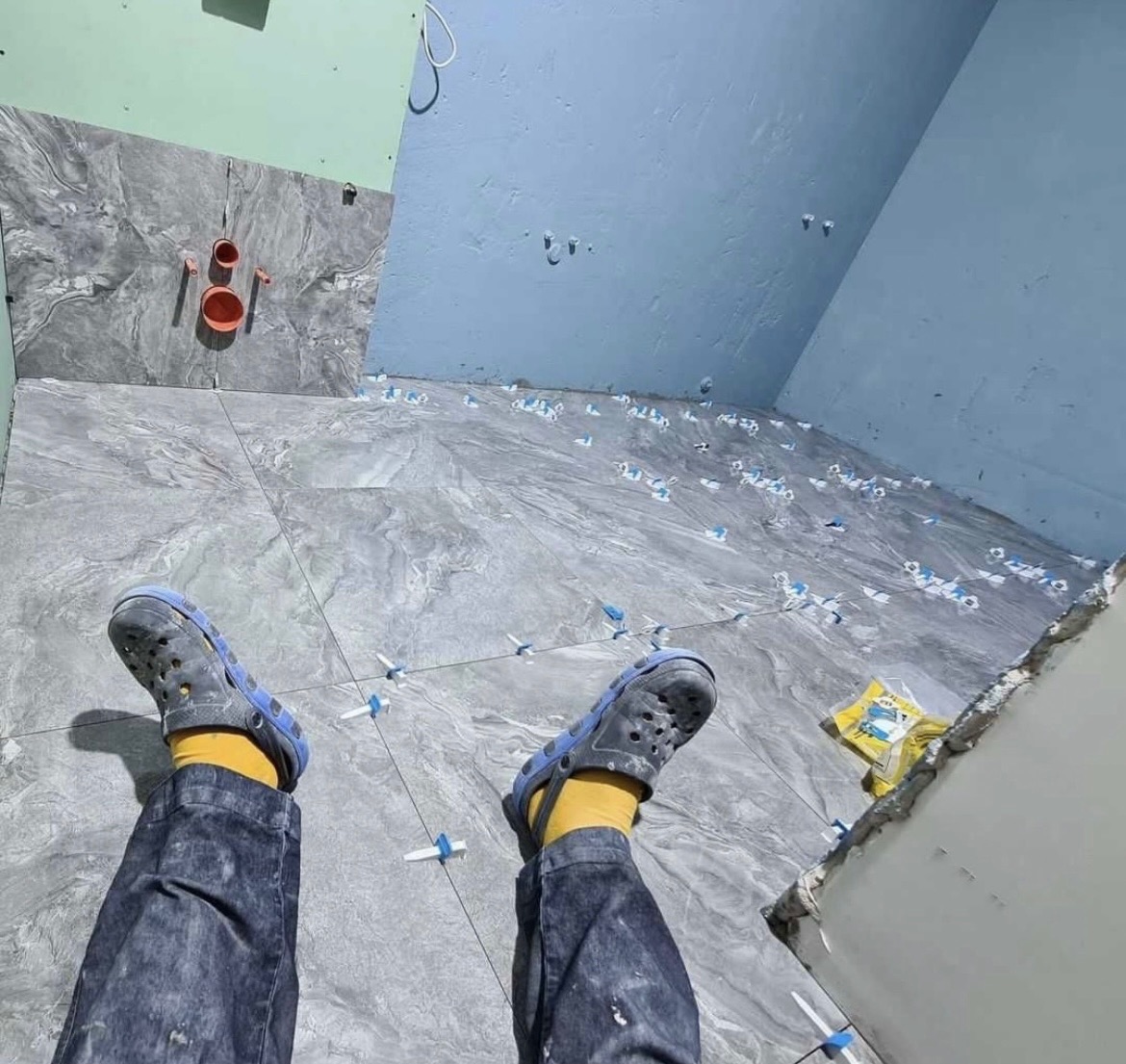 Podłoga w trakcie układania płytek imitujących marmur, widoczne krzyżyki dystansowe, rury instalacji wodnej na ścianie, niebieskie ściany i nogi wykonawcy w dżinsach i żółtych skarpetkach...