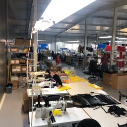 D.O.K.T.O.R. JACEK ZYGAJ SPÓŁKA KOMANDYTOWA - Wnętrze hali produkcyjnej z kilkoma stanowiskami pracy; kobieta w okularach szyje na maszynie żółty materiał przy oświetleniu lampą; na stole leżą czarne elementy z napisem 'WATERPROOF'; w tle...