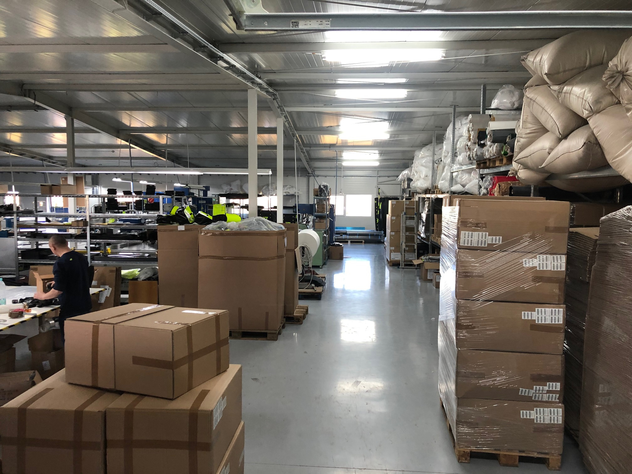 Wnętrze hali produkcyjnej szwalni z zapakowanymi kartonami, belami materiałów na półkach i pracownikiem przy stole w tle.