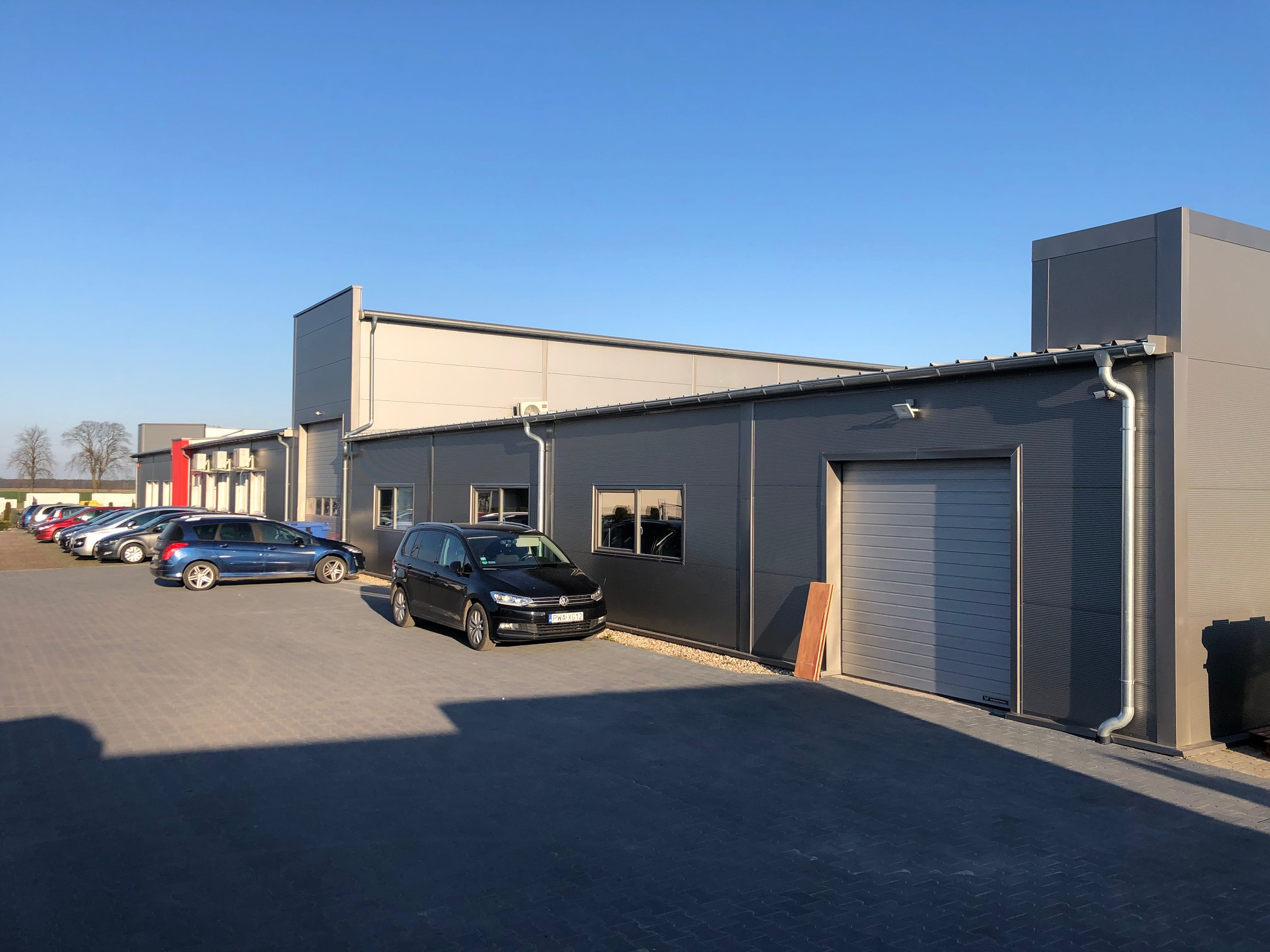 Budynek szwalni odzieży z zaparkowanymi samochodami na zewnątrz, widok na fasadę z bramą garażową i oknami.