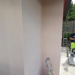 Świeżo nałożona, jasna struktura elewacyjna na rogu budynku, z widoczną drabiną, wiadrem z farbą i fragmentem pracownika w odblaskowym ubraniu w tle.