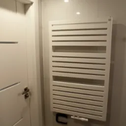 Biały, grzejnik łazienkowy drabinkowy zamontowany na jasnej, kafelkowanej ścianie w łazience. Obok widoczny fragment białych drzwi z metalową klamką.