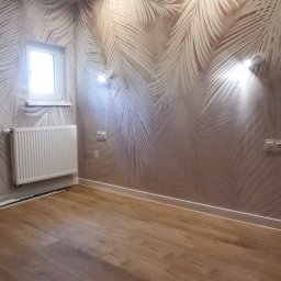 DARBUD Usługi Remontowo-Budowlane Dariusz Paradowski - Jasne panele podłogowe w pokoju z tapetą w palmy i białym grzejnikiem. Widoczne gniazdka i lampy ścienne.