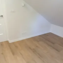 Jasne panele podłogowe w pokoju na poddaszu z białymi ścianami, widoczne gniazdka i włączniki, białe listwy przypodłogowe, drzwi z lewej strony kadru.