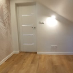 DARBUD Usługi Remontowo-Budowlane Dariusz Paradowski - Wykończone poddasze ze świeżo ułożonymi panelami podłogowymi, białą ścianą z tapetą w liście i nowoczesnymi, białymi drzwiami z poziomymi frezami.