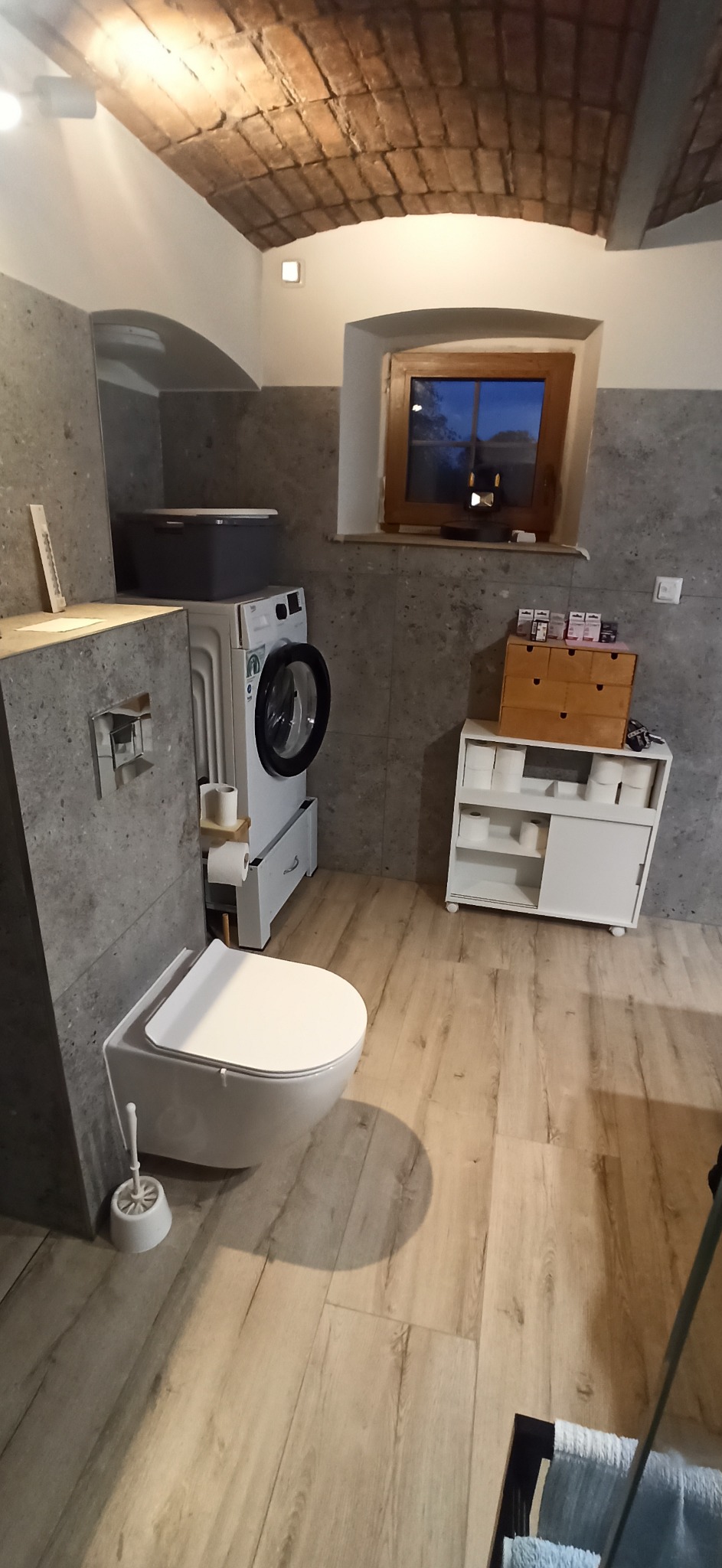 Nowoczesna łazienka z toaletą wiszącą, pralką i ceglanym sklepieniem, wykończona w szarościach i beżach.