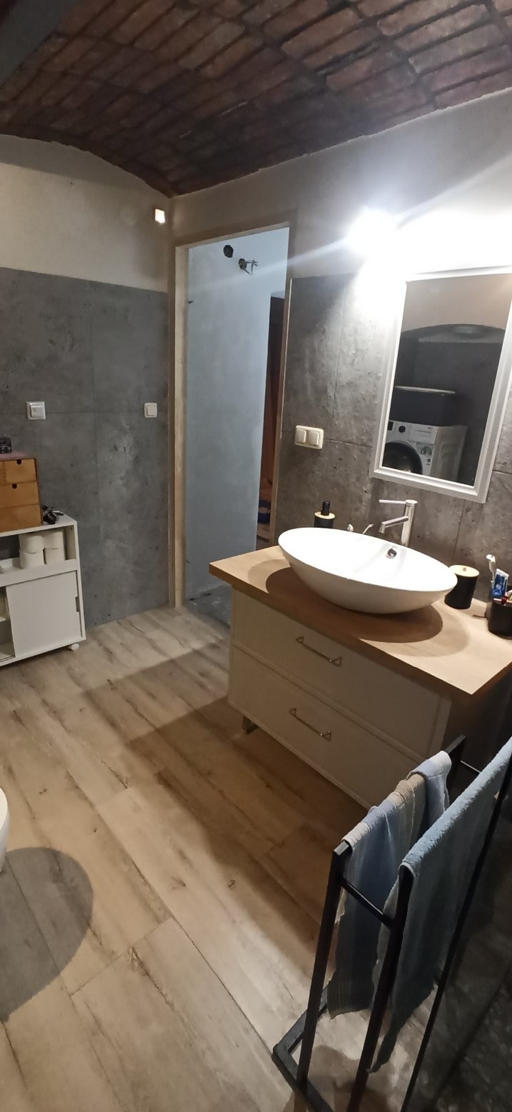 Nowoczesna łazienka z umywalką nablatową, drewnianym blatem i szarą ścianą imitującą beton. Widoczne panele podłogowe, ceglany sufit i pralka odbijająca się w lustrze.
