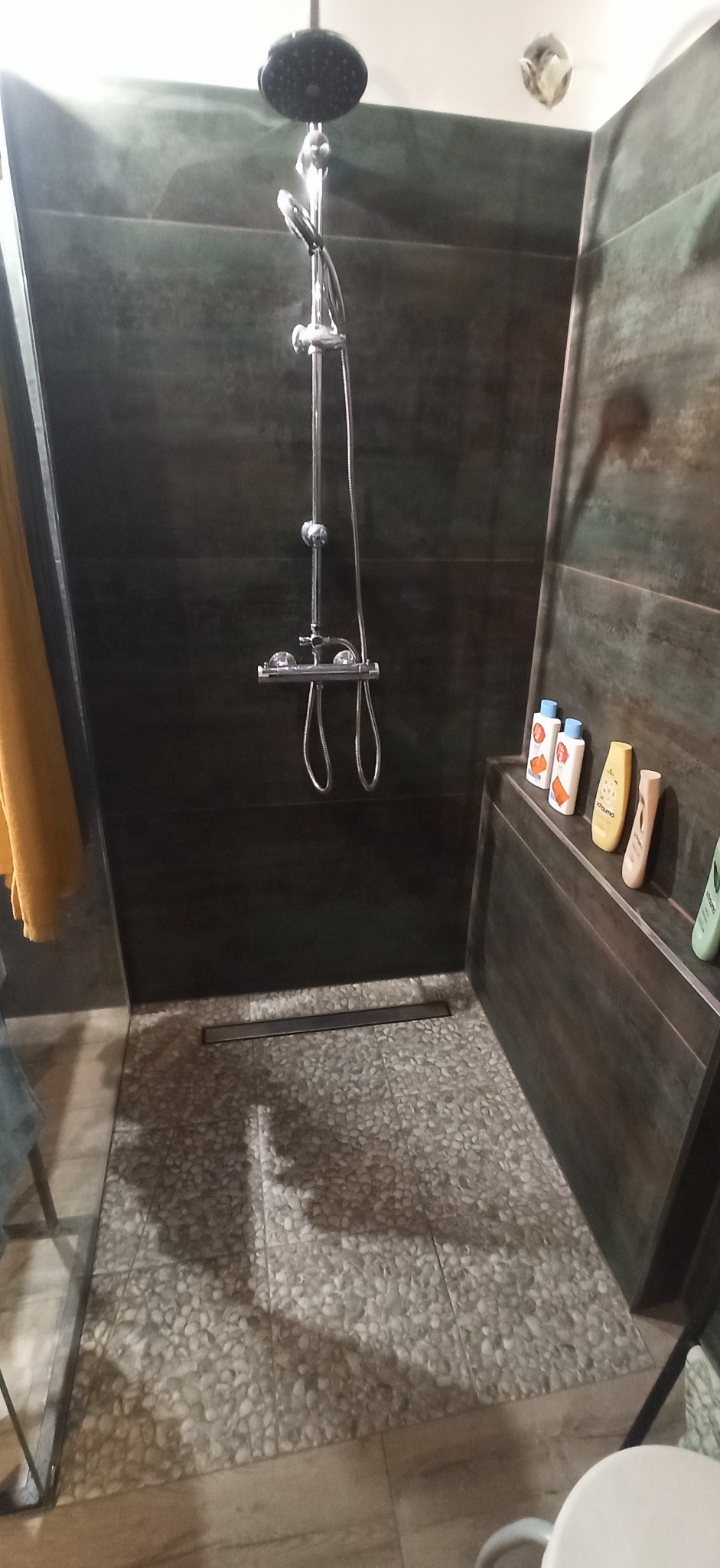 Nowoczesna kabina prysznicowa z ciemnymi, płytkami imitującymi drewno na ścianach, podłogą wyłożoną drobnymi kamykami i chromowaną armaturą. Na półce przy ścianie ustawione butelki z kosmetykami.