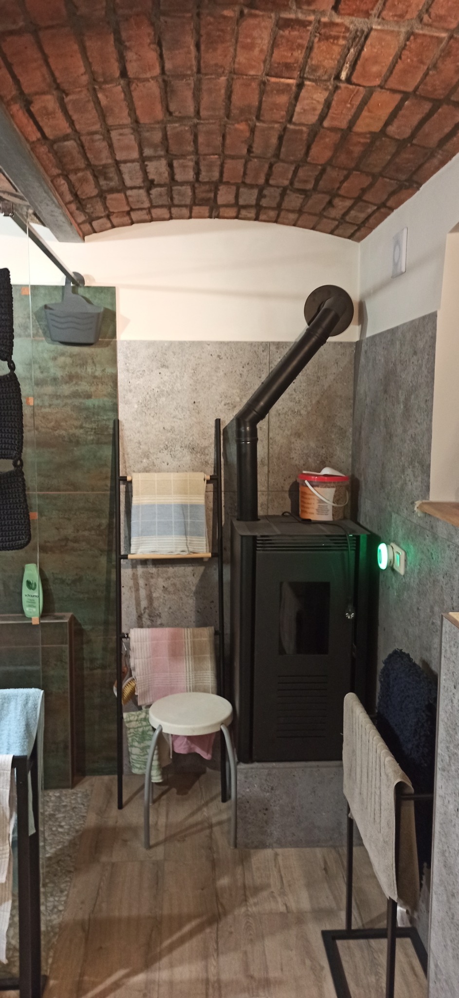 Wnętrze łazienki z ceglanym sklepieniem, piecem na drewno z czarną rurą odprowadzającą dym, drabinką na ręczniki i małym stołkiem.