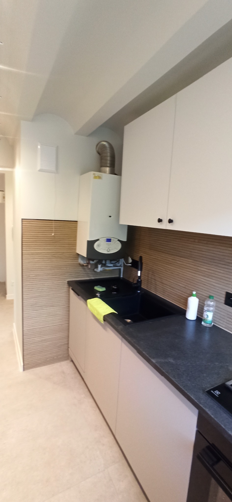 Wykończona kuchnia z kremowymi szafkami, czarnym blatem i zlewem. Na ścianie beżowe panele i biały piec gazowy. Minimalistyczny styl, nowoczesne oświetlenie.