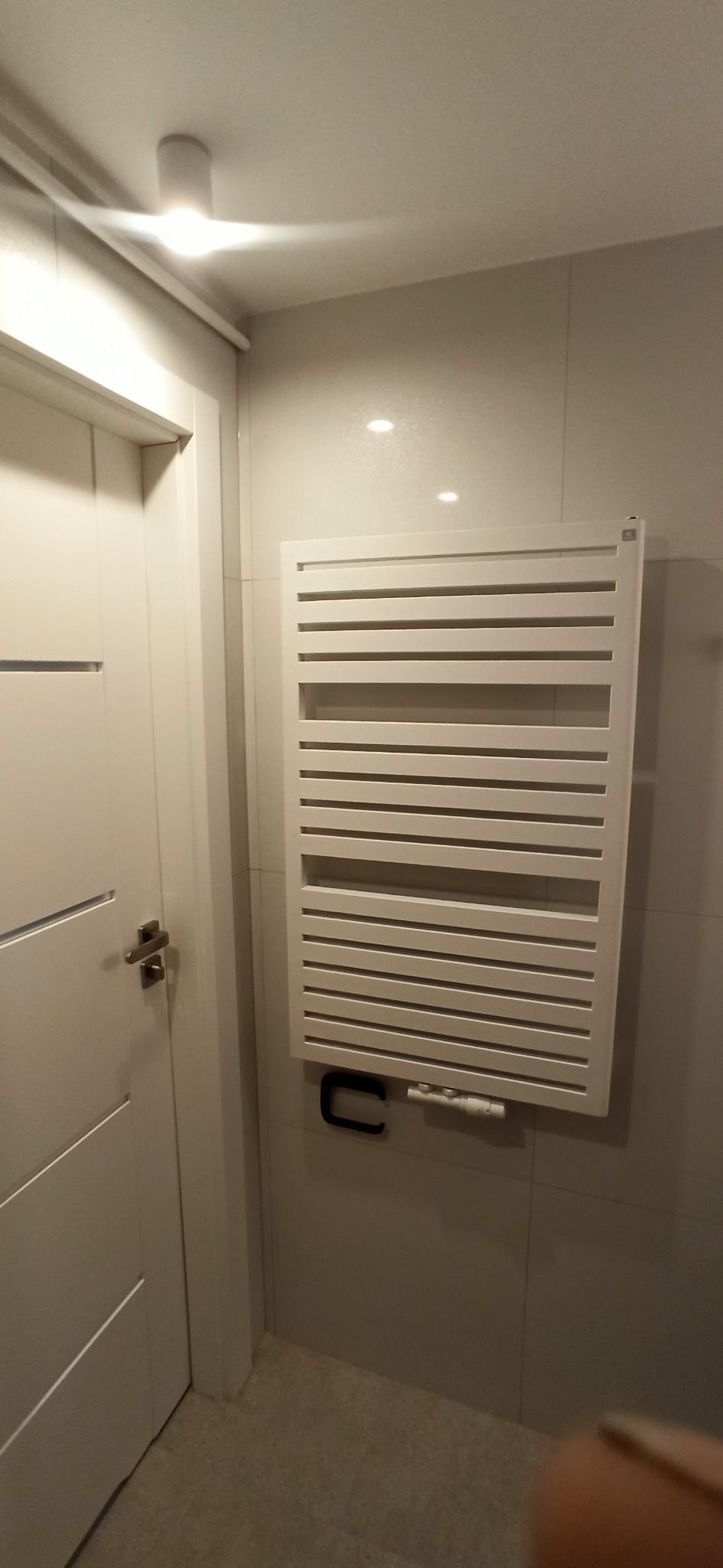 Biały, grzejnik łazienkowy drabinkowy zamontowany na jasnej, kafelkowanej ścianie w łazience. Obok widoczny fragment białych drzwi z metalową klamką.