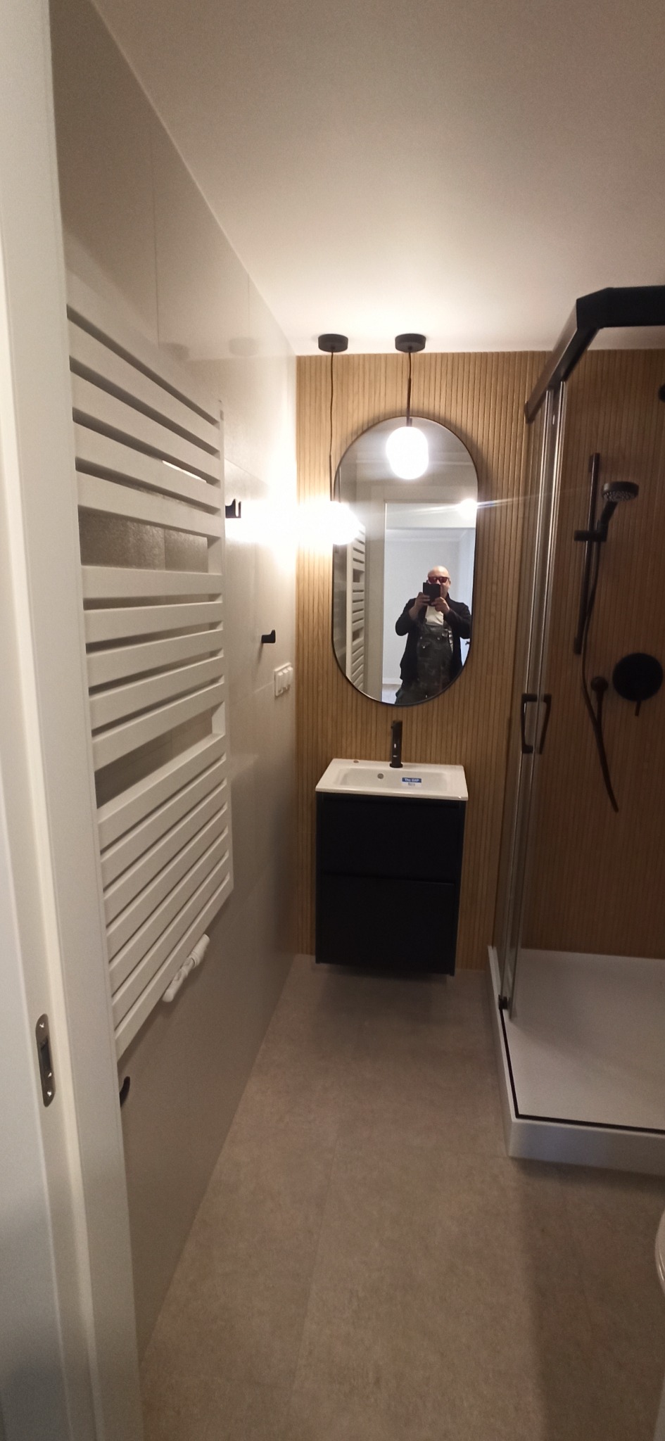 Nowoczesna łazienka po remoncie z kabiną prysznicową, umywalką z szafką i grzejnikiem drabinkowym. Na ścianie drewnopodobne panele i owalne lustro.