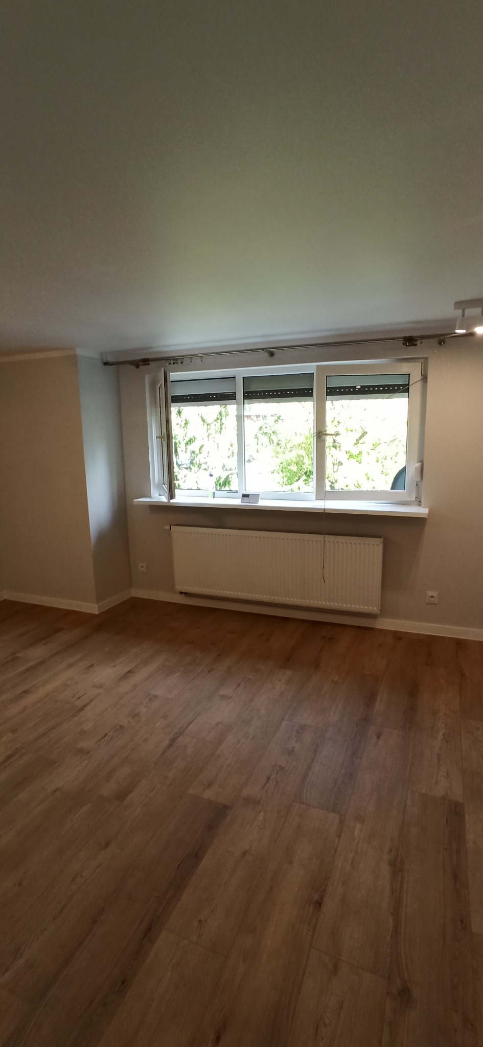 Jasne panele podłogowe w pokoju z grzejnikiem pod oknem, jedno okno otwarte. Ściany pomalowane na jasny odcień beżu. Widok na zieleń za oknem.