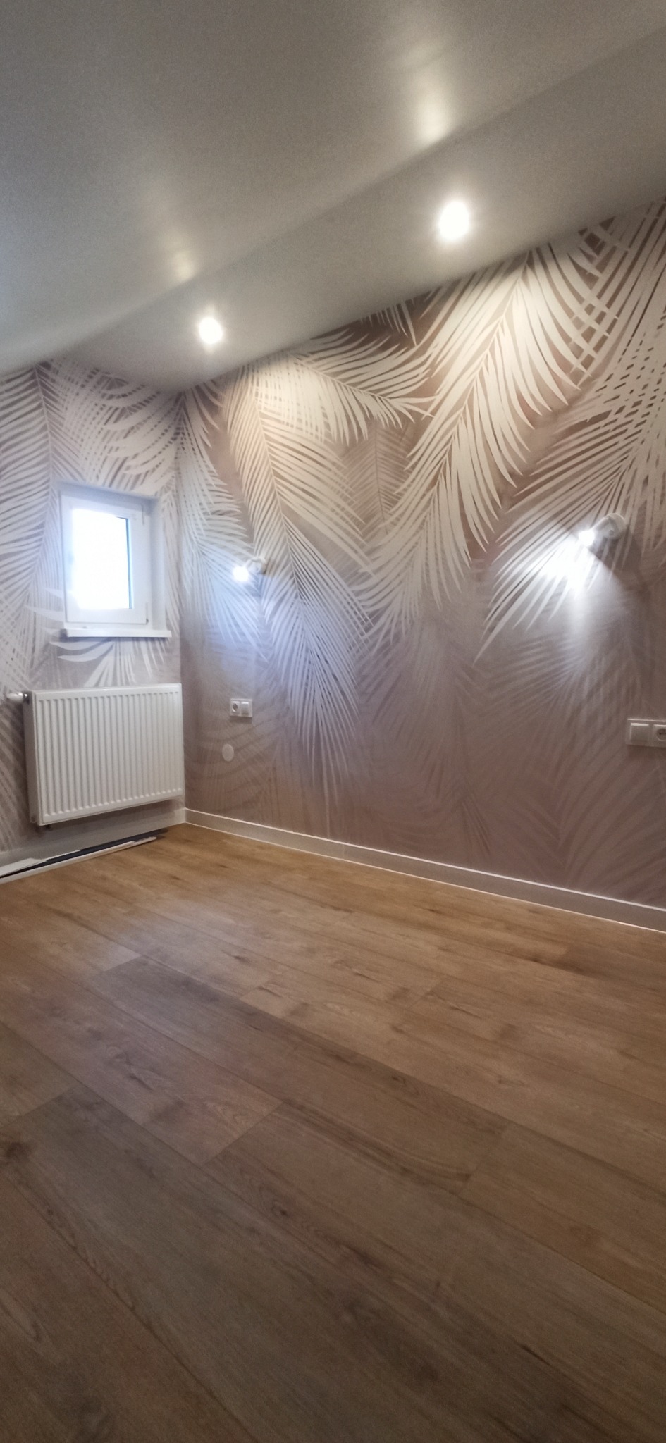 Jasne panele podłogowe w pokoju z tapetą w palmy i białym grzejnikiem. Widoczne gniazdka i lampy ścienne.