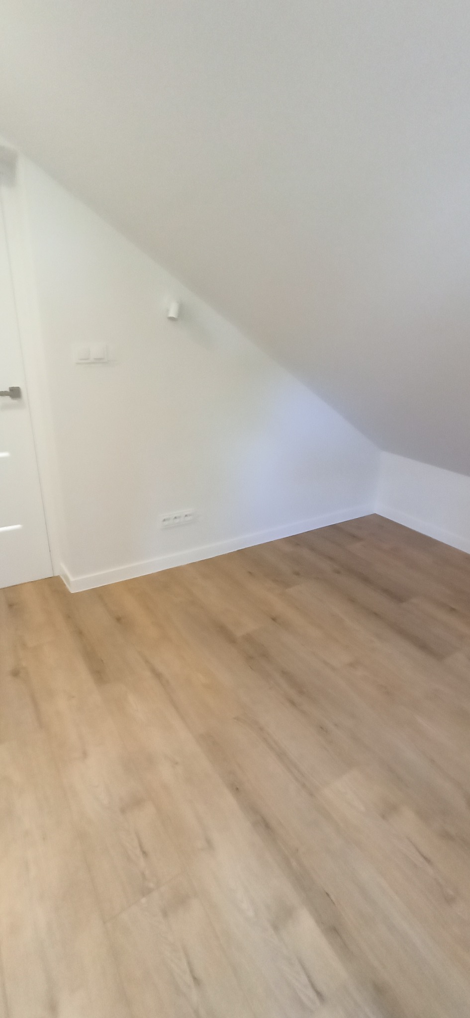 Jasne panele podłogowe w pokoju na poddaszu z białymi ścianami, widoczne gniazdka i włączniki, białe listwy przypodłogowe, drzwi z lewej strony kadru.