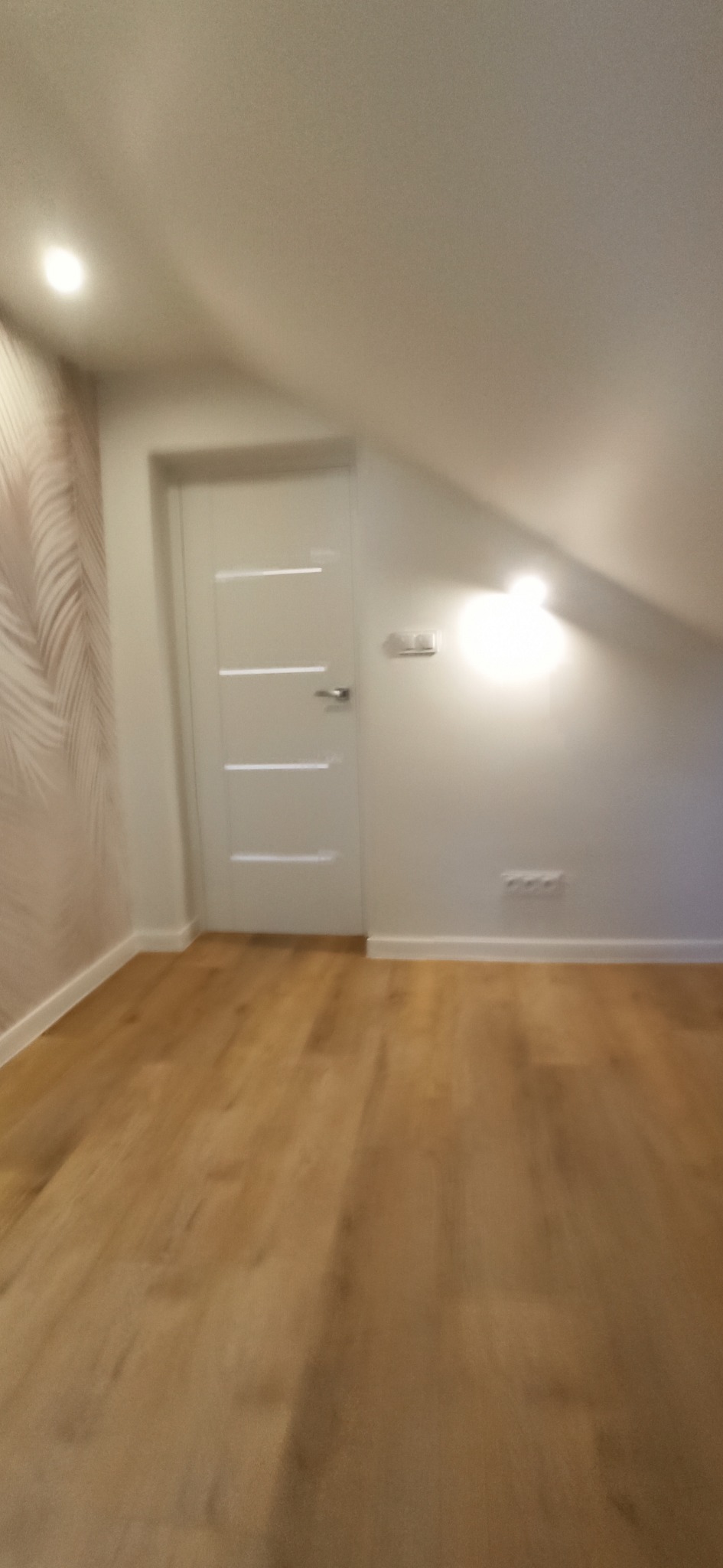 Wykończone poddasze ze świeżo ułożonymi panelami podłogowymi, białą ścianą z tapetą w liście i nowoczesnymi, białymi drzwiami z poziomymi frezami.